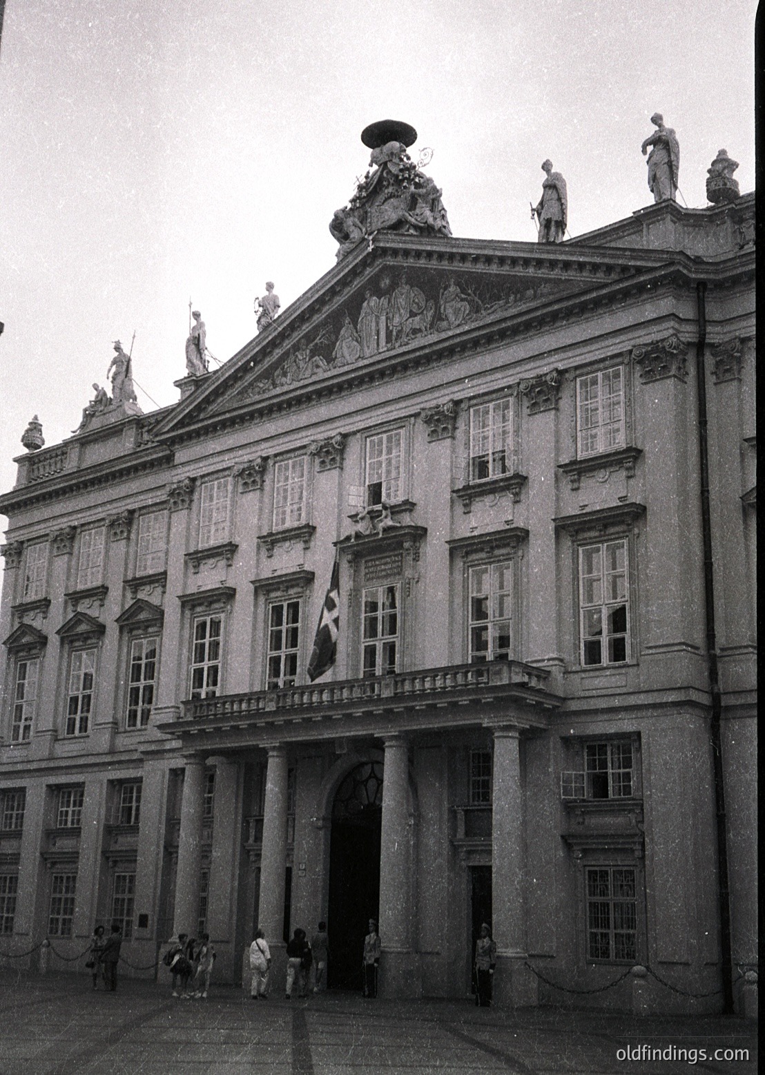 Neoclassical façade featuring symmetrical design, tall columns, and sculptural reliefs. Central archway flanked by statues atop balustrade. Mid-20th century flagpole with emblematic flag. Small crowd in courtyard, likely 1960s–1980s.