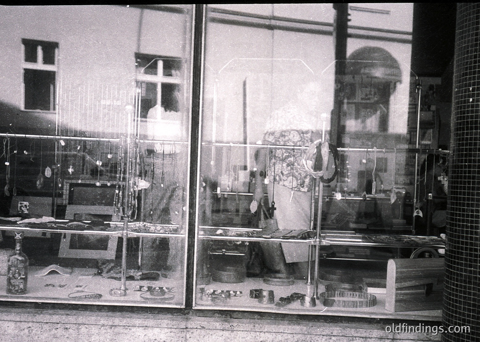 Vintage black-and-white shot of a cluttered, multi-tiered display window showcasing household goods—pottery, textiles, and metalware. Reflections reveal a building with arched windows and a dome. Likely Eastern European, mid-20th century.