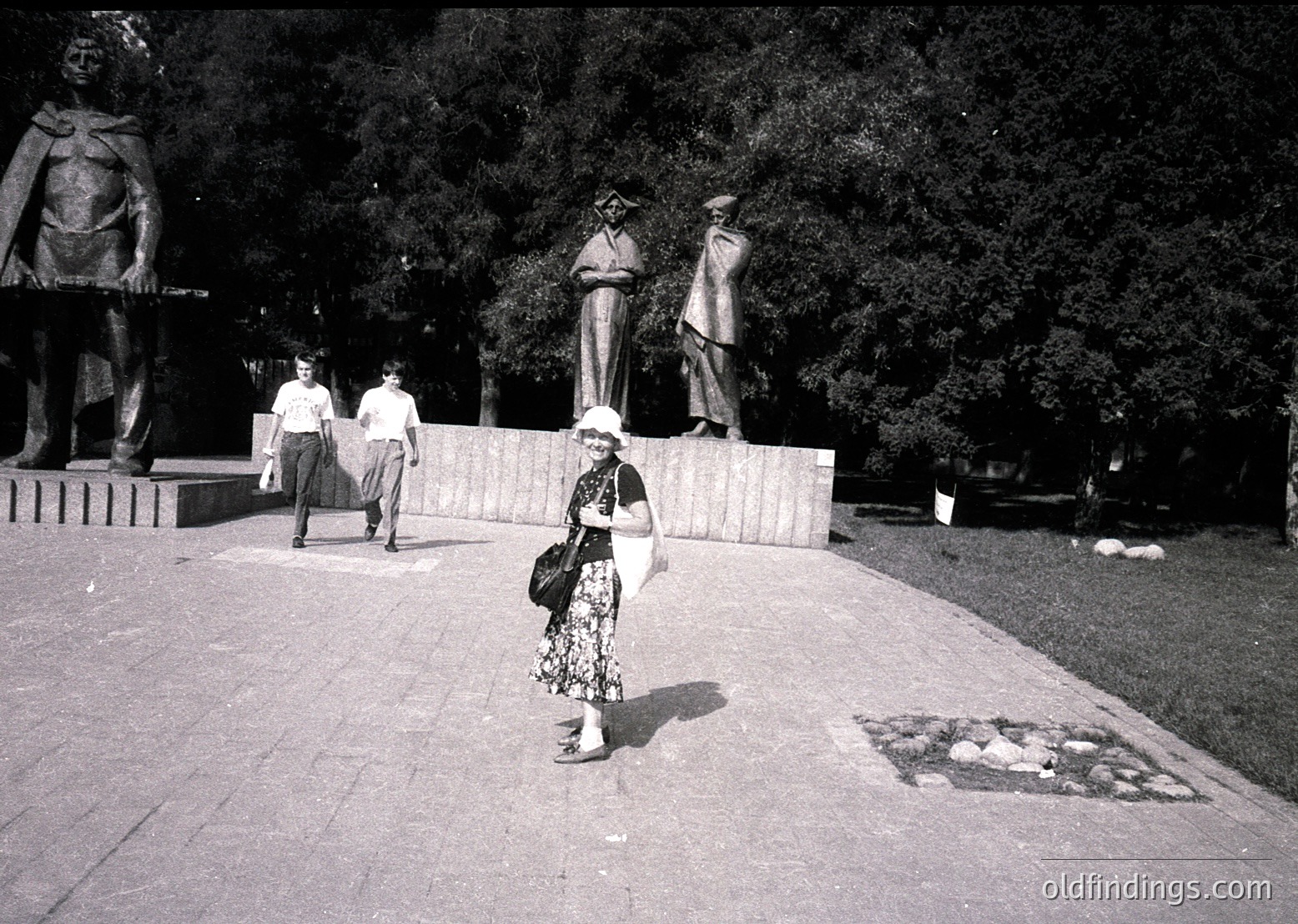 Black-and-white street scene featuring three prominent bronze statues of laborers in mid-gesture atop a raised platform, likely depicting industrial or agricultural themes. A woman in a floral dress and hat carries a handbag, walking toward the camera on a paved plaza. Trees and additional pedestrians frame the scene, suggesting a public square or park. Mid-20th century urban design with Soviet-era influences.
