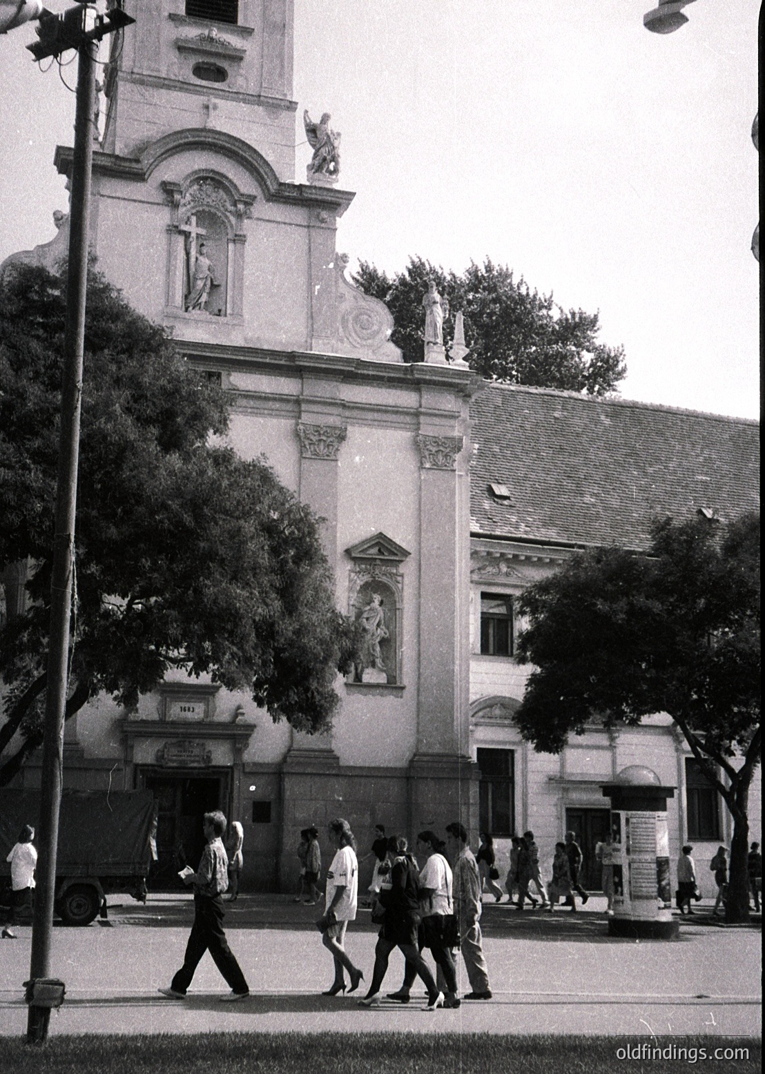 Neoclassical building façade with sculpted reliefs and a bell tower, likely from the early 20th century. Group of casually dressed pedestrians crossing a street in front. Urban setting with mature trees and vintage street lamps.