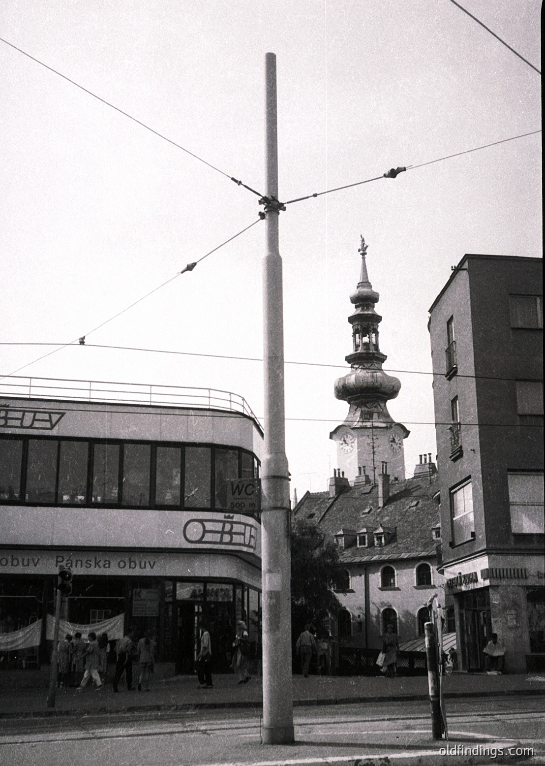Mid-20th century urban street corner blending Soviet-era and historic architecture. Prominent tram pole and overhead wires dominate foreground. Left: "ČSOB" bank and "Panská obuv" (shoe store) signage in Art Deco-style building. Right: Baroque-style church tower with onion dome and spire. Pedestrians in 1960s-era clothing.
