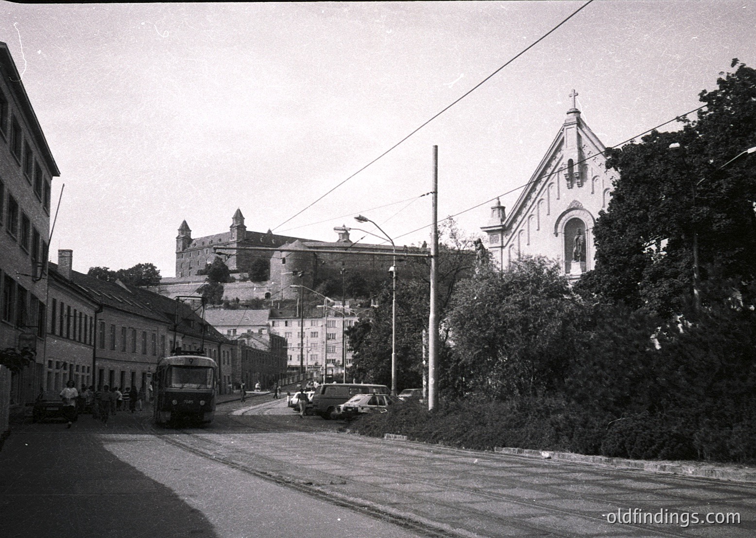 Black-and-white street scene featuring a vintage bus and cars on a gently curving road, flanked by mid-20th century European architecture. Prominent Gothic-style church with twin spires and a fortress-like castle perched on a hill in the background. Dense greenery and utility poles line the right side. Likely Eastern Europe, 1950s–1960s.
