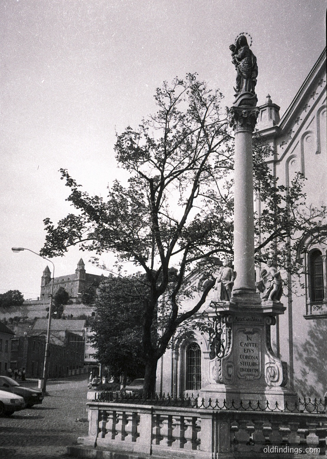 Historic stone fountain with Baroque-style statue of Virgin Mary and child atop, flanked by cherubs. Engraved plaque reads *"In Carpe di Corona Stella"* (likely 18th–19th c.). Urban setting with castle ruins in background, suggesting European heritage.