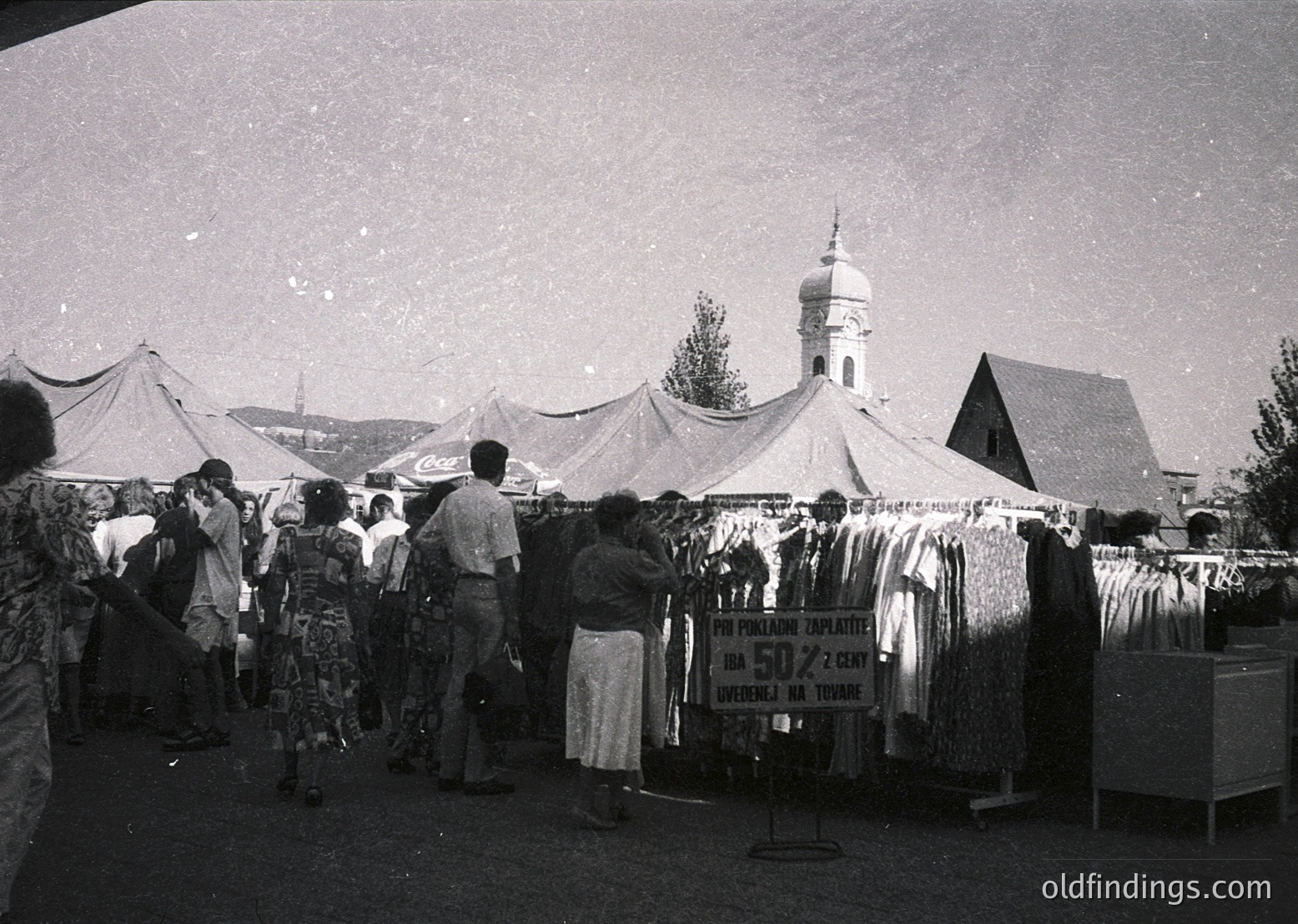 Outdoor market scene with clothing stalls under white tents, featuring a sign in Cyrillic advertising 50% discounts. A domed church with a cross and traditional wooden structures in background. Mid-20th century Eastern European setting, likely Bulgaria.