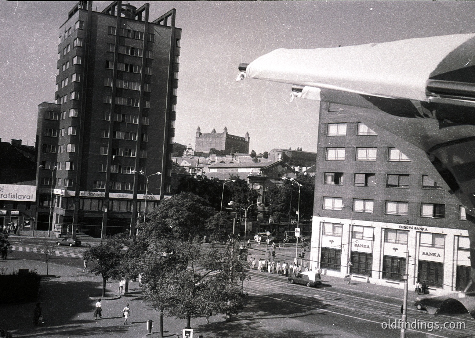 Mid-century urban scene featuring Brutalist architecture: concrete high-rises with exposed formwork, a prominent "Hotel Bratislava" sign, and a "Spoja" building. Historic castle perched on a hill in background. Pedestrians and cars populate a wide, tree-lined boulevard. --- *(Note: Approximate time period inferred from architectural style and signage; no direct date visible.)*