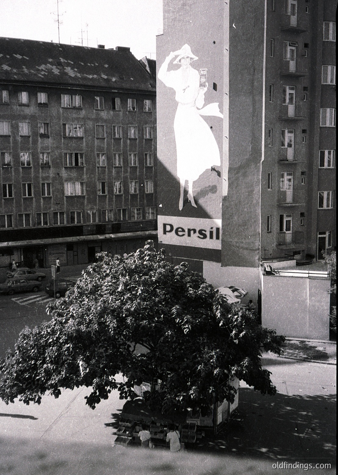 Vintage 1960s-70s urban billboard featuring a stylized white silhouette of a woman in a flowing dress, advertising **Persil** detergent. The poster dominates a building facade in a European city, with multi-story residential blocks and a tree partially obscuring the lower frame. Urban architecture and mid-century advertising design evident.