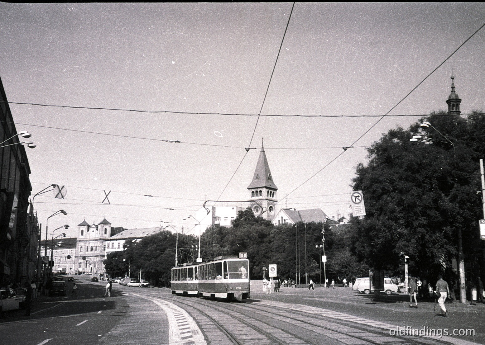 Classic mid-20th century European street scene with vintage tram on tracks. Prominent church steeple and historic buildings flank the road, suggesting urban core. Overhead wires and street signs (, ) indicate public transit infrastructure. Pedestrians and parked cars add to urban atmosphere.