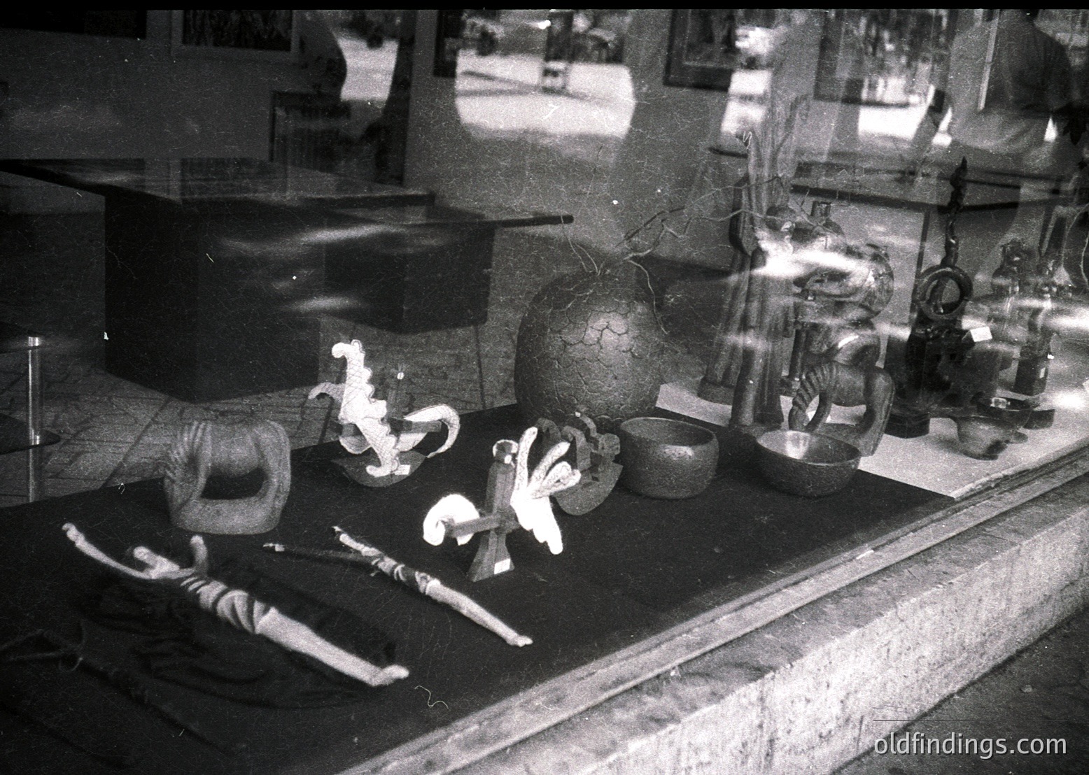 Vintage black-and-white display of vintage bicycles and ceramic items in a shop window. Two bicycles in mid-air, one with a rider in a dynamic pose, alongside spherical and cylindrical pottery. Reflections and blurred figures suggest a bustling street scene. Likely or European setting.