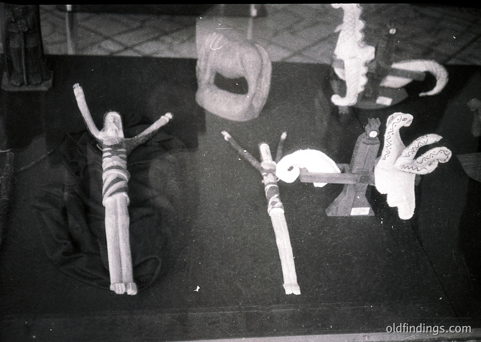 Vintage black-and-white photo of two wooden dolls in a crucifixion pose, flanked by a small ceramic bird and a decorative object resembling a stylized fish. Reflections and shadows suggest a glass display case. Likely 1950s–1970s European folk art or religious craft.