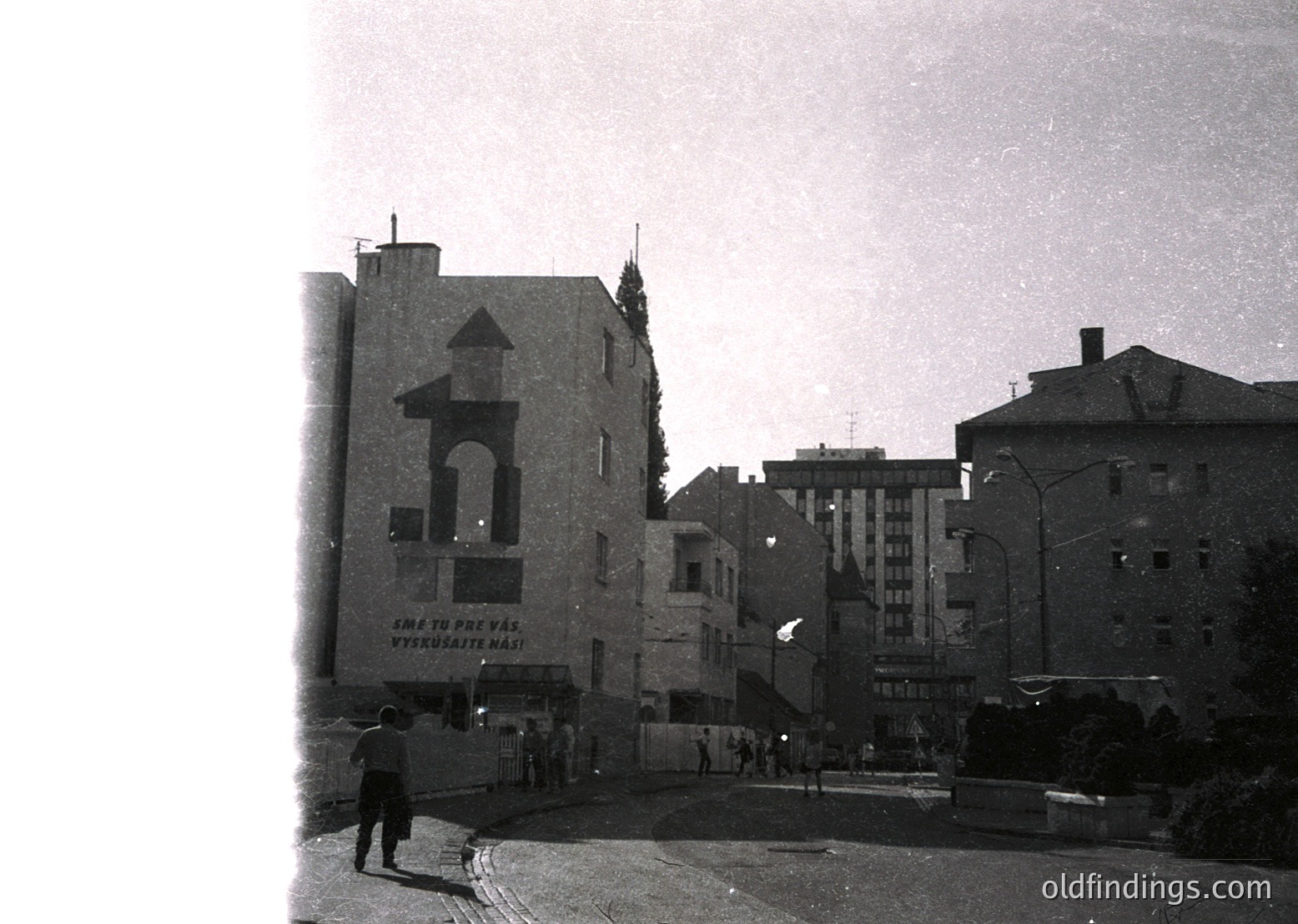 Mid-20th century urban street corner featuring a mix of Soviet-era architecture. Prominent building with Cyrillic signage reading *"Еме то прева" / "Вероятно на"*, likely Bulgarian. Pedestrian crossing empty street with vintage streetlamps and minimal traffic. Residential/apartment blocks in background.