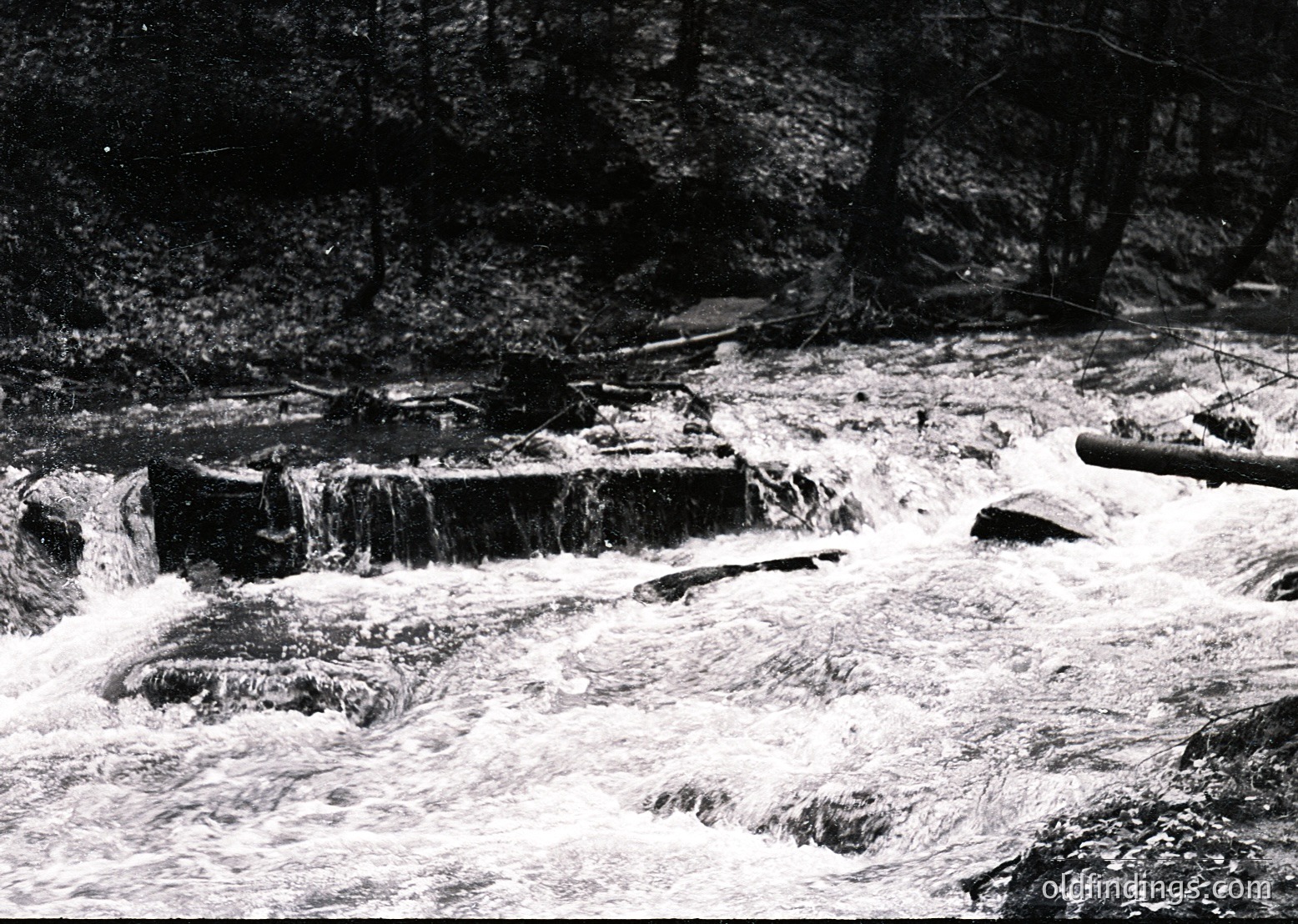 Black-and-white shot of a cascading waterfall with concrete steps guiding flow through a forested ravine. Vegetation frames both sides, with a wooden bridge or platform visible upstream. Likely mid-20th century due to monochrome and styling.