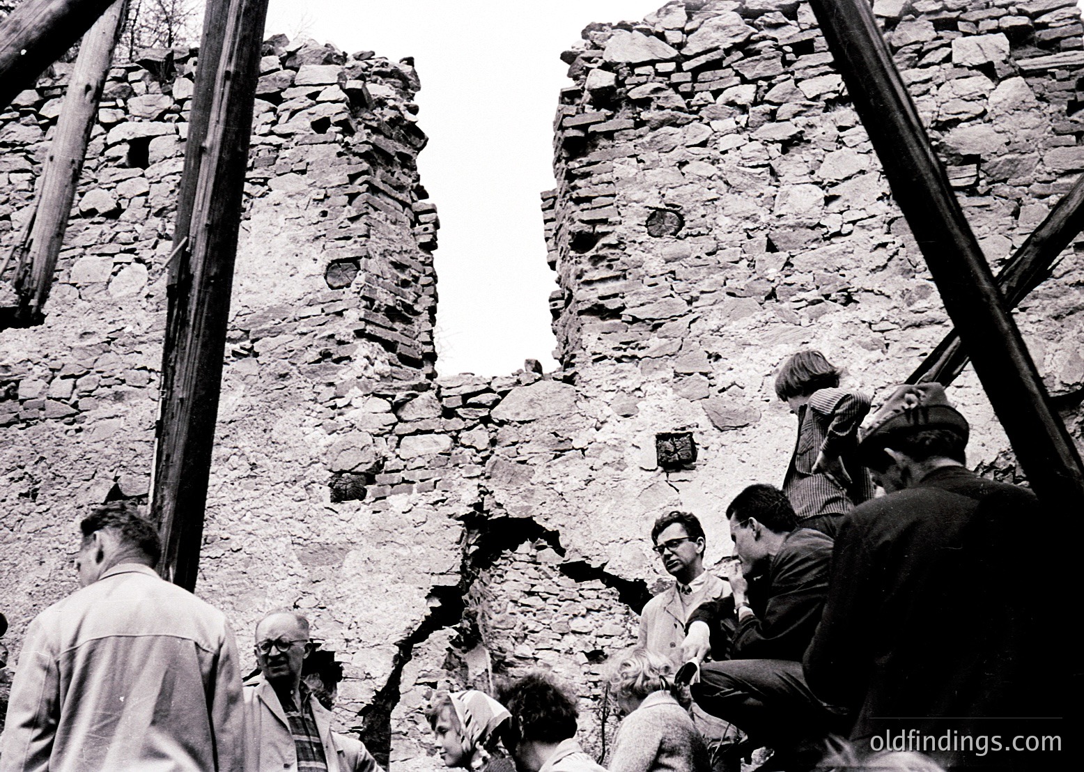 Black-and-white photo of a group inspecting a damaged stone wall, likely post-earthquake or collapse. Timber supports stabilize the structure. Mid-20th century clothing suggests or . Urban or historical site with rough-hewn stonework.