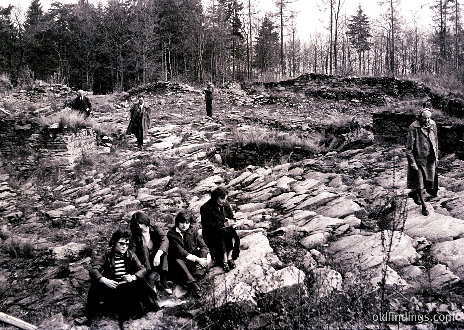 Black-and-white photo of a group exploring a rocky archaeological site in a forested area, likely Eastern Europe, 1960s-70s. Four seated individuals examine ruins, while others stand or walk nearby. Distinctive stone walls and uneven terrain suggest a historic or prehistoric settlement.