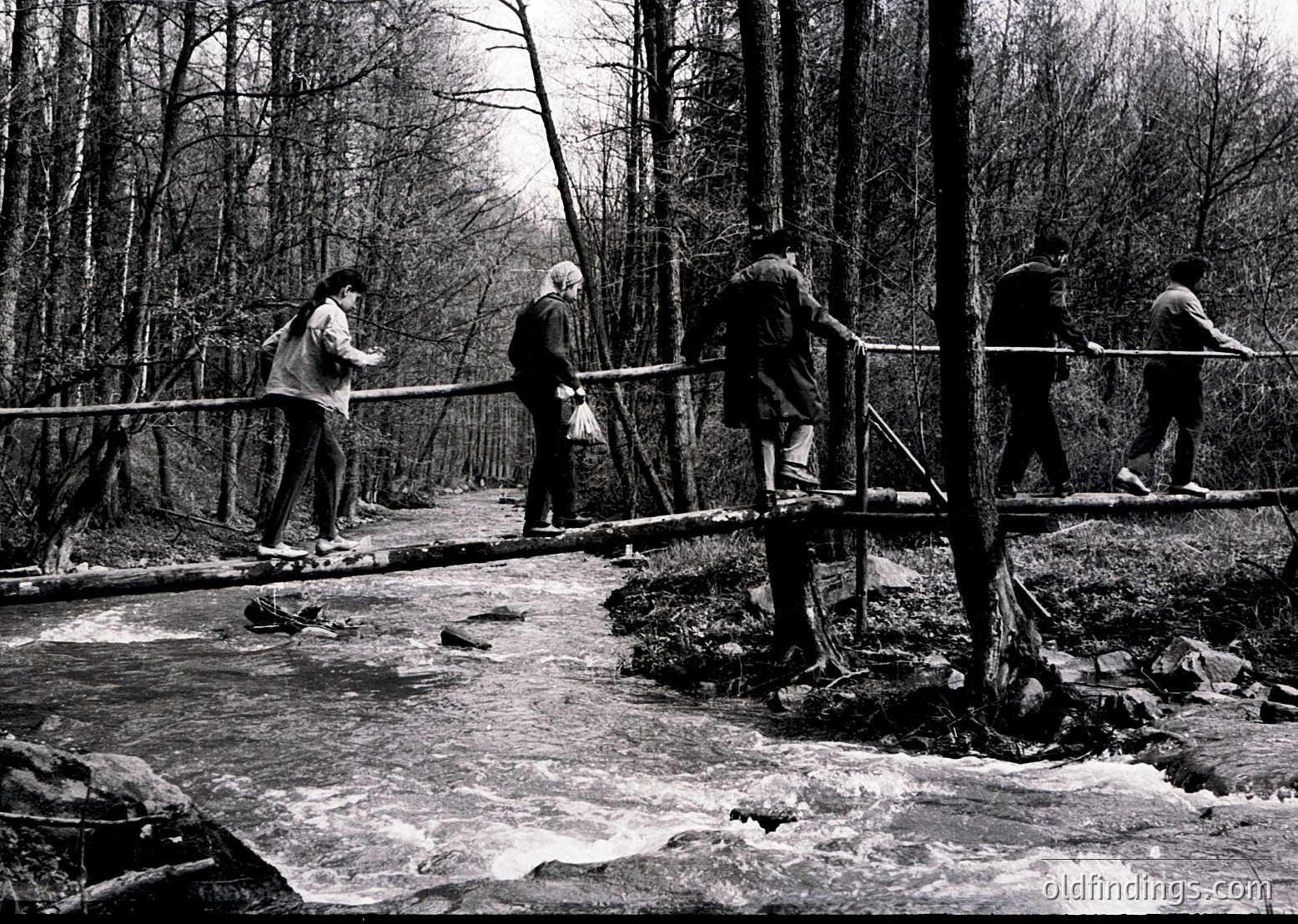 Four individuals cross a wooden suspension bridge over a shallow, rocky stream in a forested area, likely mid-20th century. Bridge handrails and bare trees suggest autumn/winter.