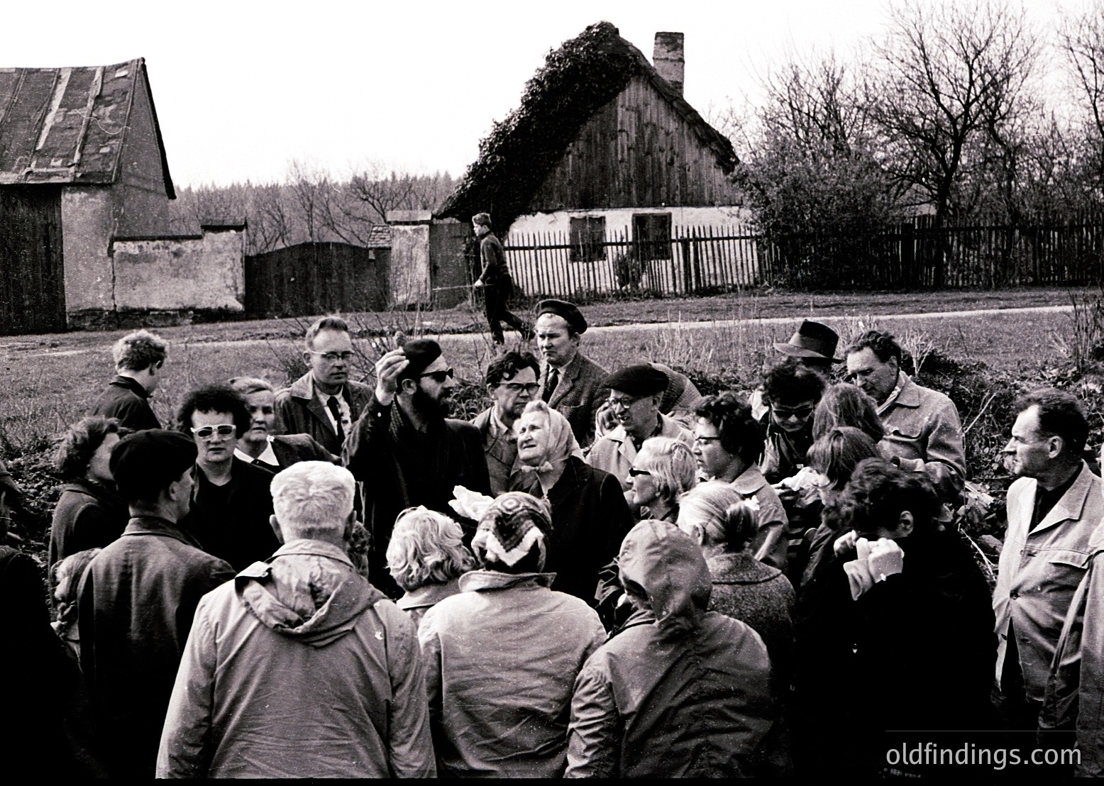 Mid-20th century rural gathering in Eastern Europe, likely –. Crowd surrounds a central speaker, possibly a local official or community leader, in a barren field near rustic wooden structures. Men and women in practical attire—overcoats, hats, and scarves—engage intently. Barren trees and a weathered fence suggest late autumn/winter. Evokes themes of