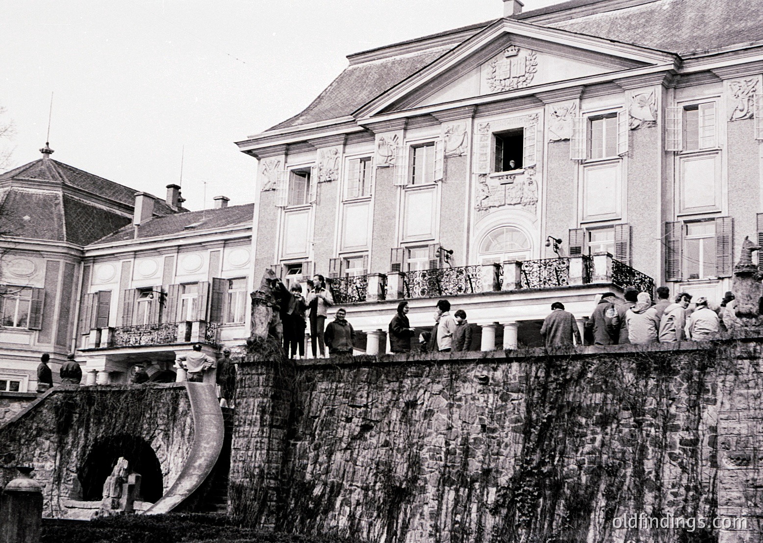 Neoclassical building with ornate balconies and arched windows, likely from the early-to-mid 20th century. Crowd of people gathered on a stone embankment and balcony, suggesting a public event or gathering. Waterway with a stone bridge arch below. [Historic European architecture with ornate balconies and arched windows, mid-20th century ]