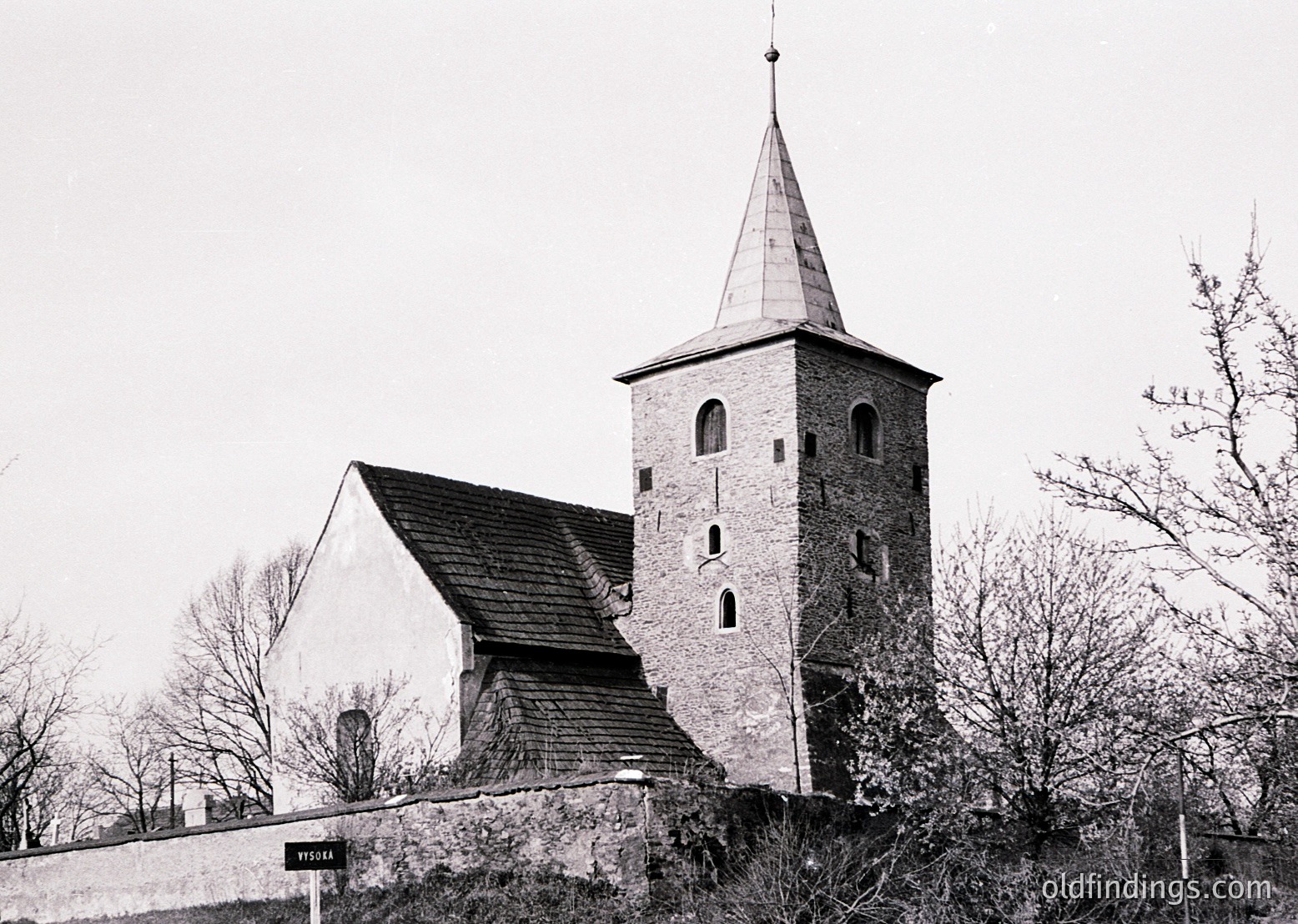 Historic stone church with pointed bell tower and steep gable roof, likely 19th-century Scandinavian architecture. Timber-framed annex visible left. Surrounded by leafless trees and a low stone wall.