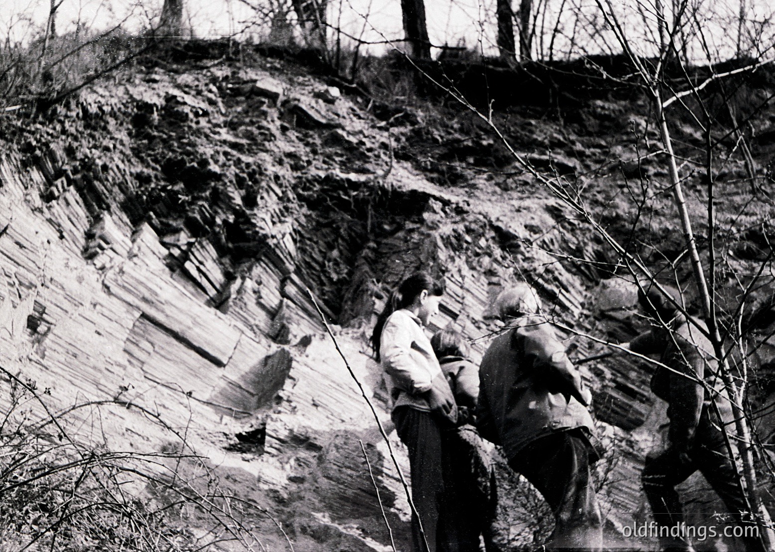 Two men examine exposed geological strata in a rocky cliffside, likely during mid-20th century. Distinct sedimentary layers and sparse winter vegetation suggest a field study or mining inspection.