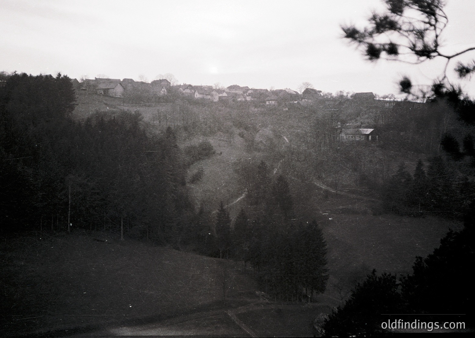Vintage black-and-white landscape featuring a rural hillside settlement with scattered houses and dense forest. Rolling terrain with winding paths and sparse vegetation suggests early-to-mid 20th century European countryside.