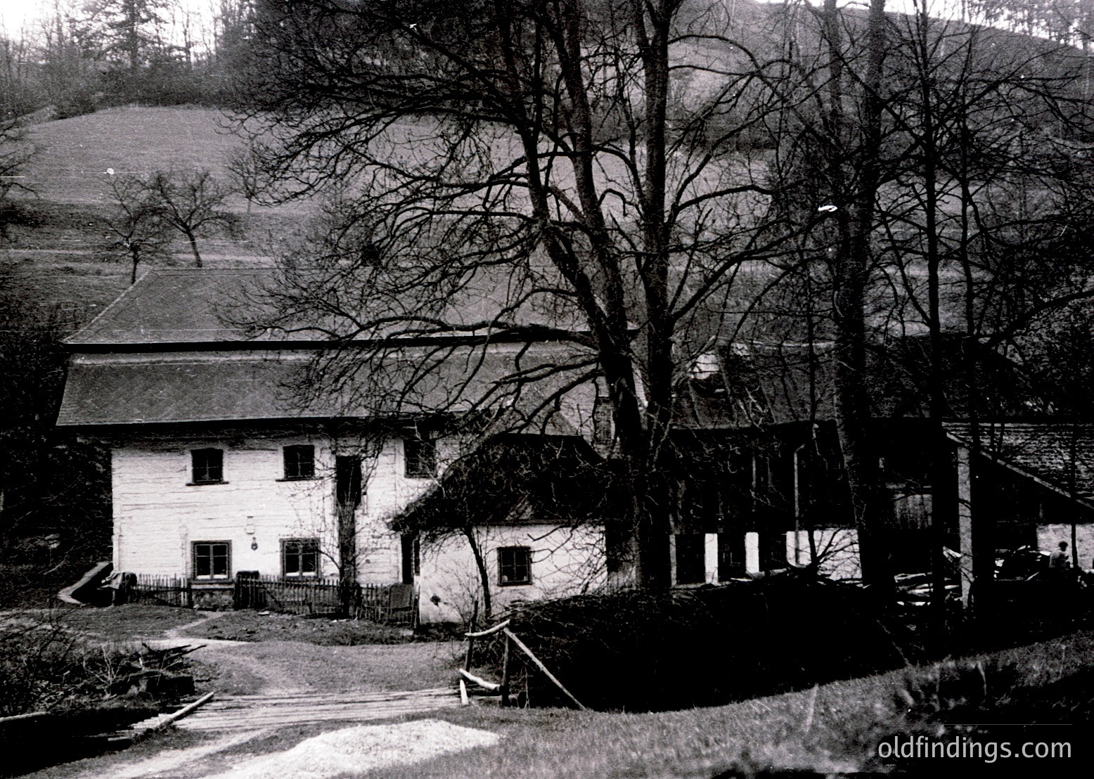 Two-story rural house with pitched roofs, surrounded by bare trees and rolling hills. Likely early-to-mid 20th century Alpine or Central European architecture.