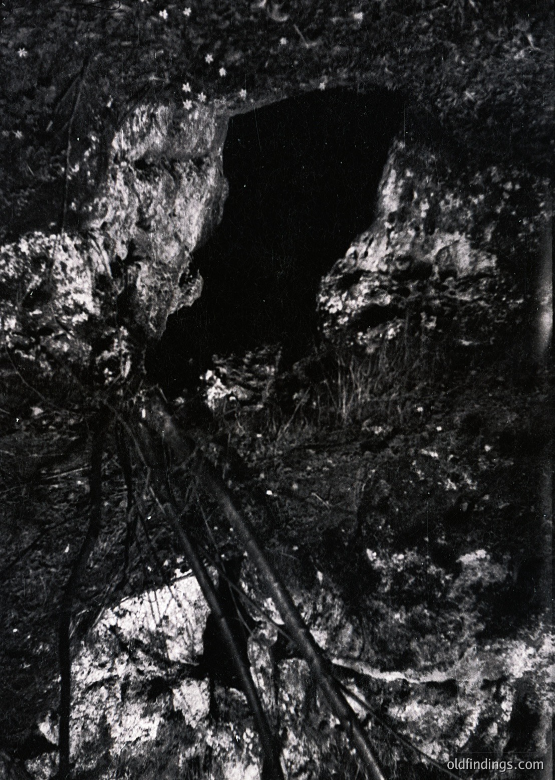 High-contrast black-and-white shot of a narrow, vertical cave entrance framed by jagged rock formations. Minimal light reveals a dark void beyond.