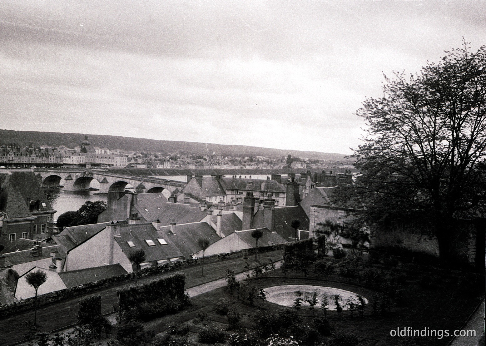 Mid-20th century European town from elevated vantage point, showcasing dense rooftops, a stone bridge, and a circular fountain in foreground. Architectural style suggests historic European urban planning. Foggy atmosphere enhances nostalgic vintage feel.