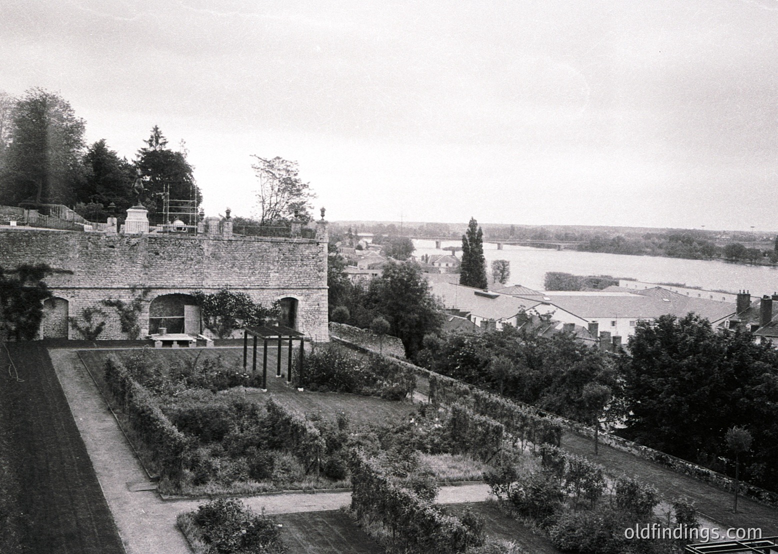 Historic estate garden with formal raised beds, stone walls, and a river valley beyond. Likely late 19th to early 20th century European countryside architecture.