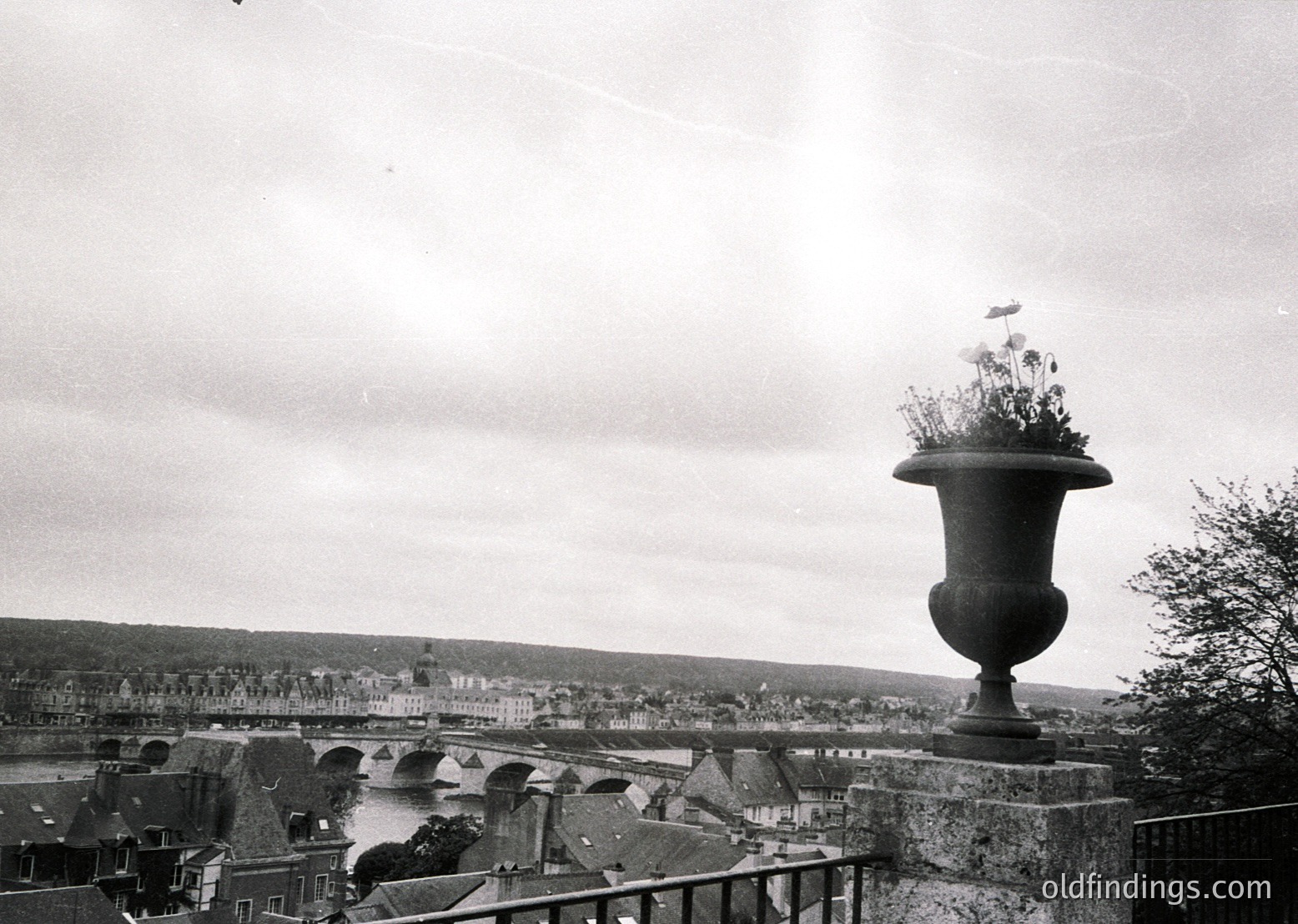 Vintage black-and-white shot of a large ornamental urn atop a stone balustrade, overlooking a river and historic town. Mid-20th century European architecture with low-slung buildings and a prominent bridge.