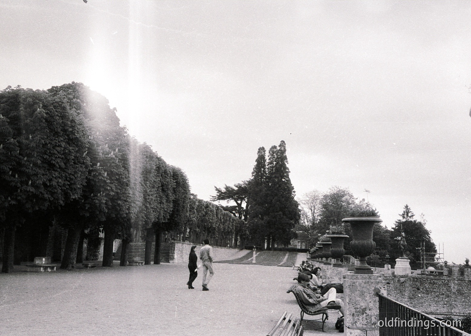 Black-and-white shot of a grand park avenue lined with mature trees and classical stone fountains. Two figures—one walking, one seated on a bench—highlight the serene, mid-20th-century atmosphere. Architectural details suggest European urban planning.
