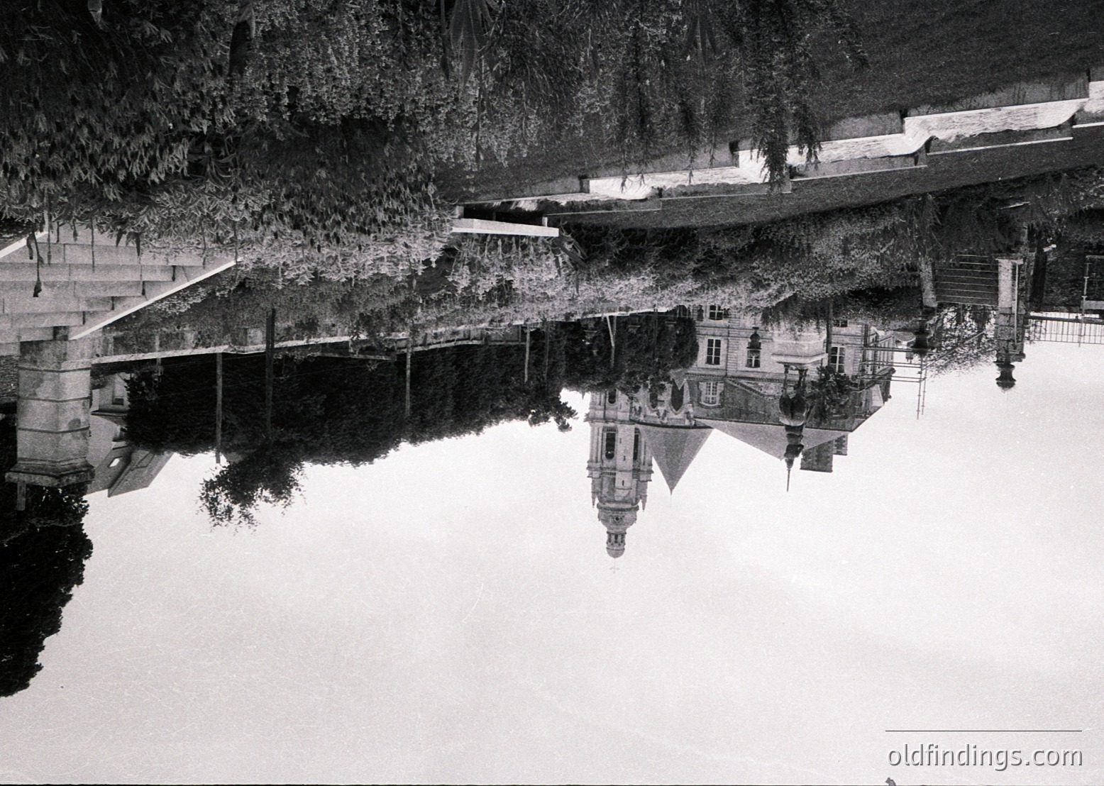 Symmetrical reflection of a grand staircase and classical building in a serene pond, framed by dense foliage. Architectural details include balustrades and arched windows. Likely European, mid-20th century.