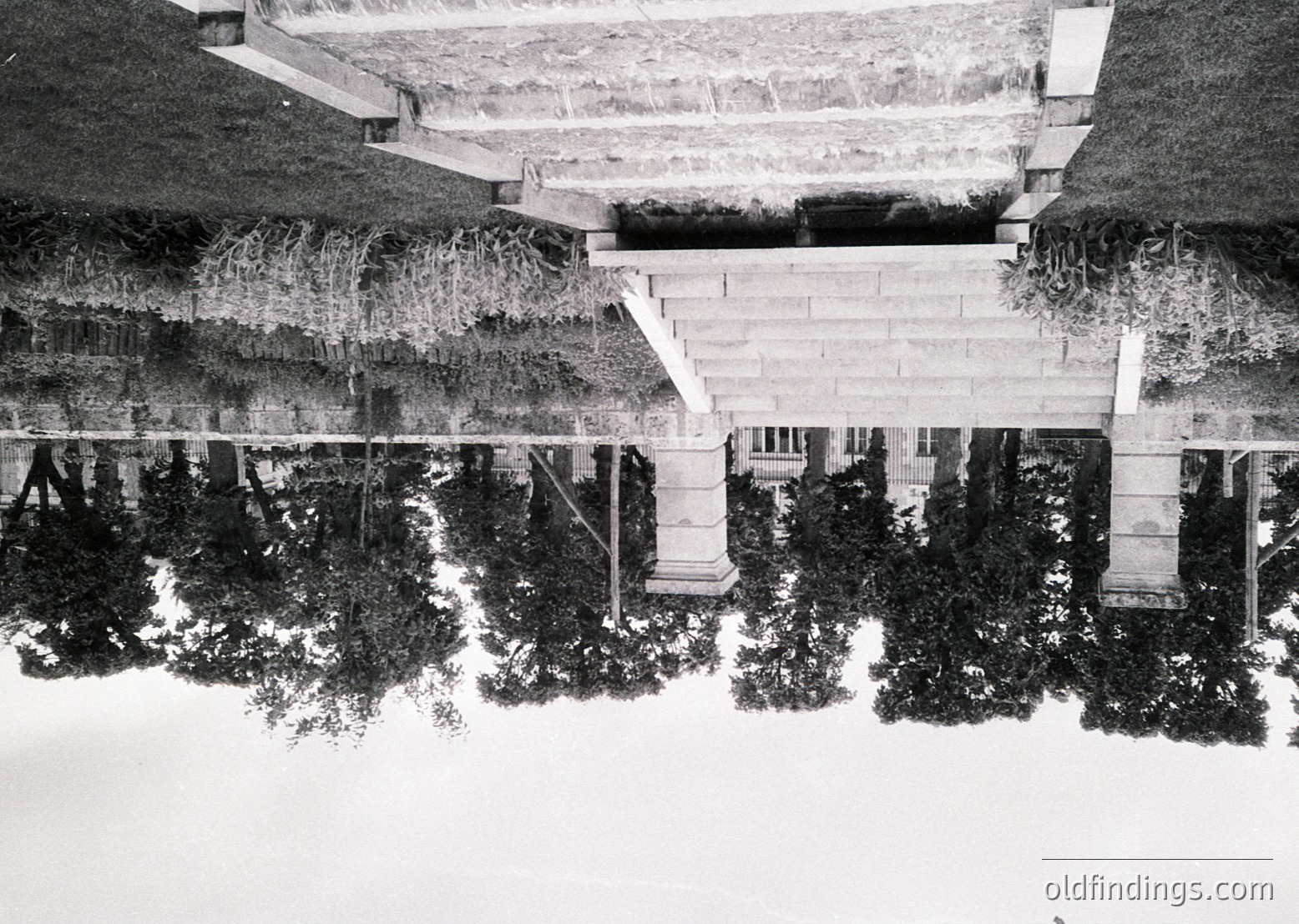 Symmetrical architectural reflection in a shallow pool, featuring classical columns and steps. Likely a 19th–early 20th century European villa or public building.