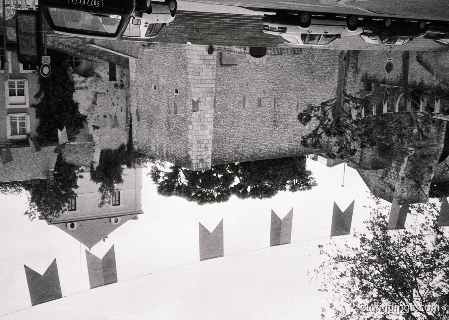Aerial view of a courtyard with mirrored water reflecting historic European architecture, likely 1960s–1970s. Symmetrical buildings with arched windows and balconies flank a central fountain. Decorative metal "M" signs line the pool’s edge, suggesting a public or institutional setting.