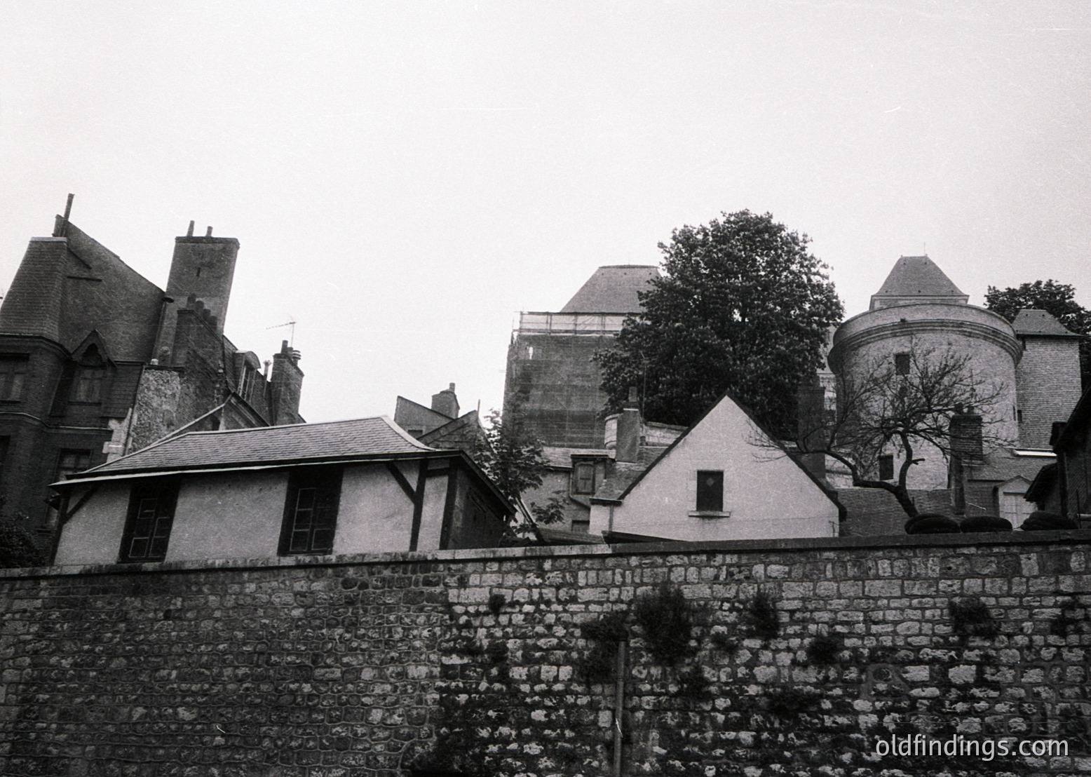 Historic European courtyard with stone walls and mixed-era architecture—19th-century brick buildings flank a central tower. Overgrown trees and a sloped roof suggest aged maintenance. Likely a castle or institutional complex.