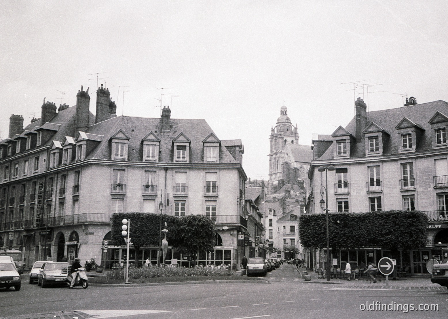 Classic European street scene featuring mid-20th century architecture. Symmetrical row of multi-story buildings with mansard roofs, dormer windows, and wrought-iron balconies. Prominent clock tower in background suggests historic town center. Vintage vehicles—including a scooter and sedan—indicate or era. Lush greenery and street lamps enhance urban charm.