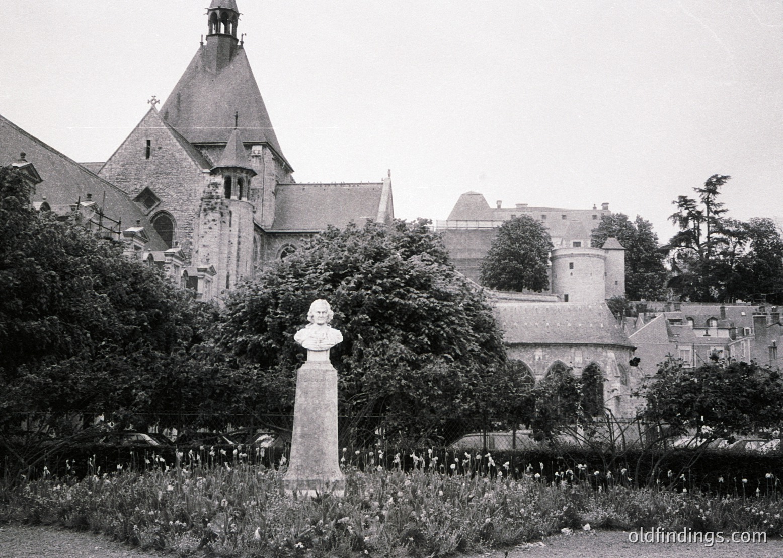 Historic black-and-white photo of a European castle courtyard with Gothic Revival architecture—steepled towers, crenellated walls, and arched gateways. Central bust statue on pedestal amid lush greenery. Likely 19th-century European estate ( ).