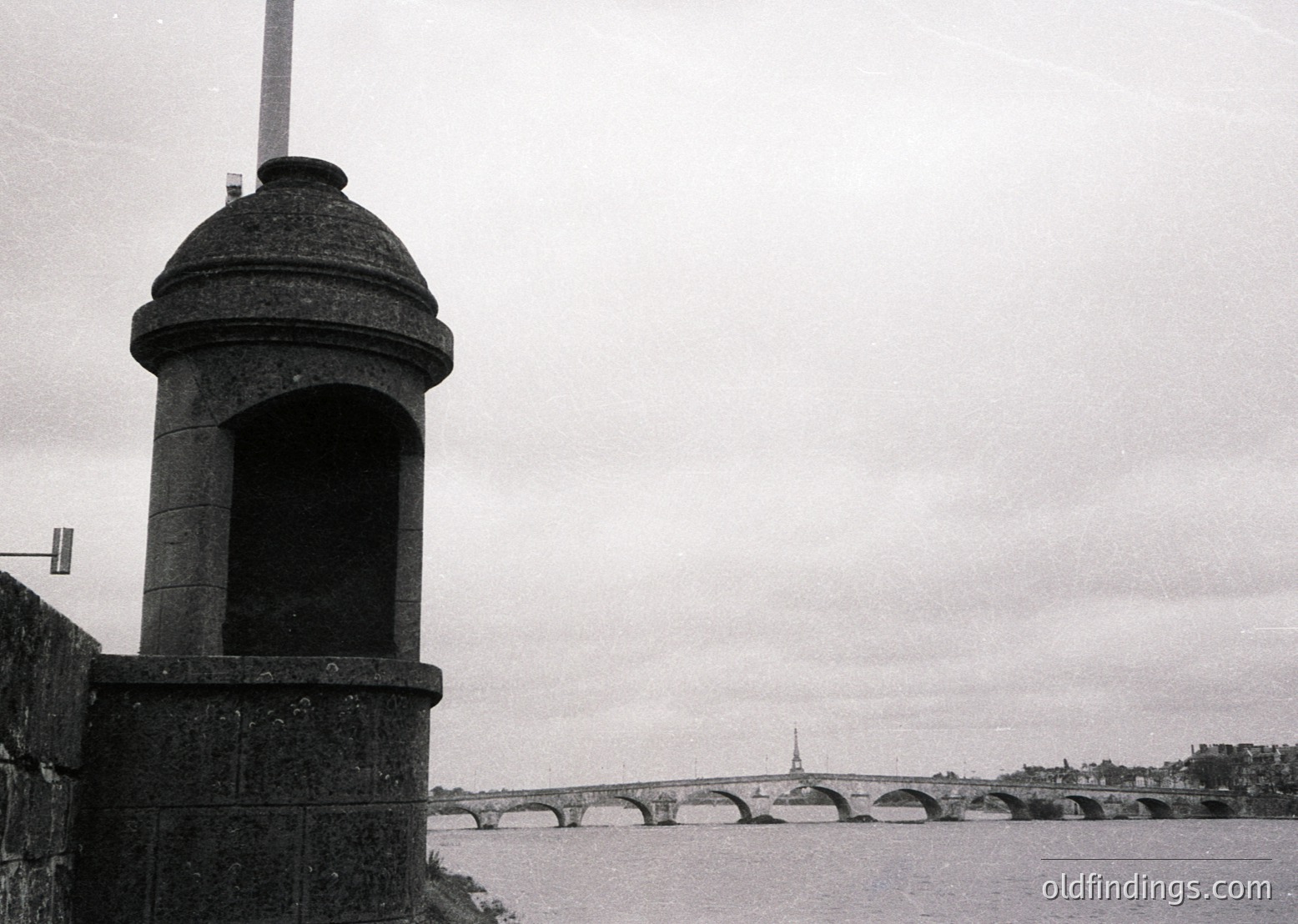 Monumental stone sentry tower with arched niche overlooking a multi-arched bridge spanning a river. Classic Parisian architecture with Eiffel Tower visible in background. Black-and-white vintage aesthetic suggests early-to-mid 20th century. Ideal for historical urban studies or retro design references.