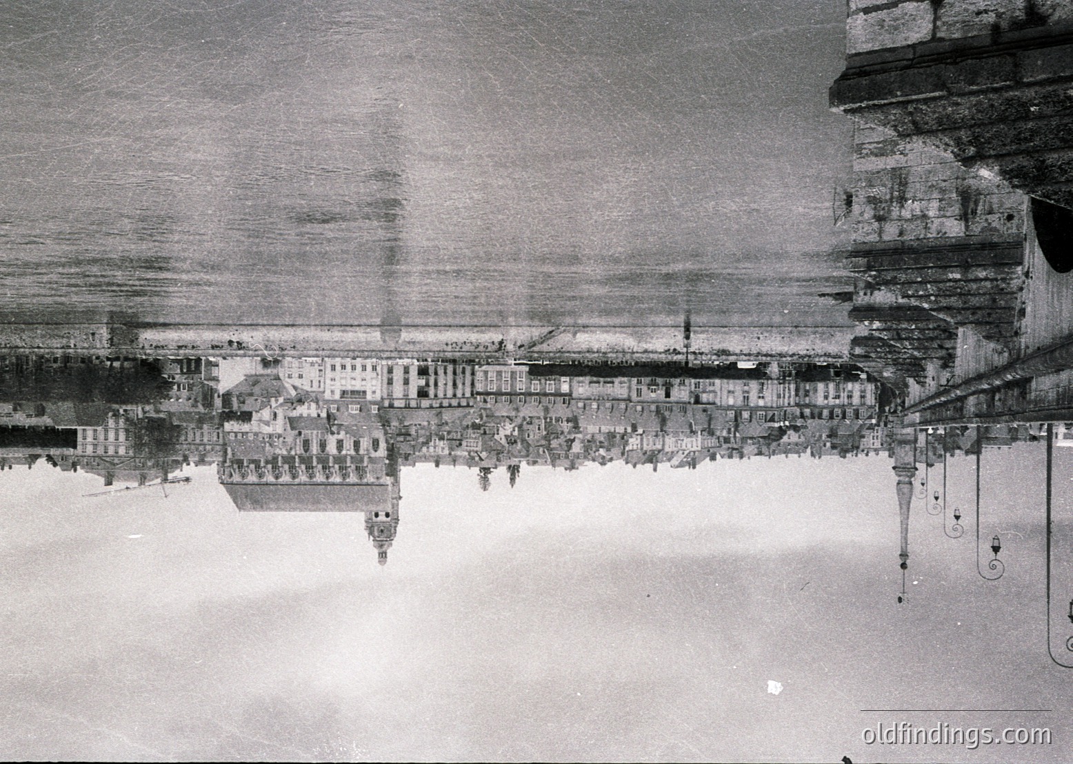 Aerial black-and-white photograph of a coastal cityscape, likely from the early-to-mid 20th century. Symmetrical row of uniform buildings along a waterfront, with a prominent pier extending into the sea. Industrial chimneys and cranes visible, suggesting port activity. Reflections in calm waters enhance composition.