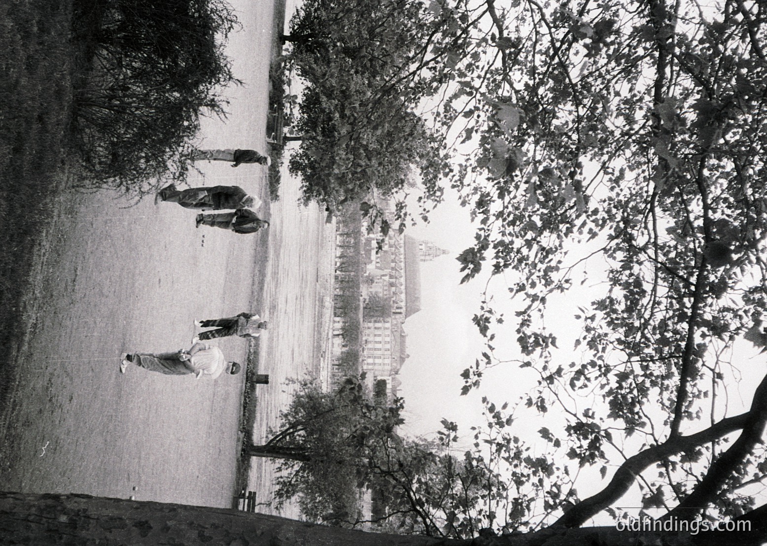 Vintage black-and-white shot of a seaside promenade with three figures in mid-stride, framed by leafy trees. The curved concrete wall and distant beach huts suggest a mid-20th-century European coastal resort.