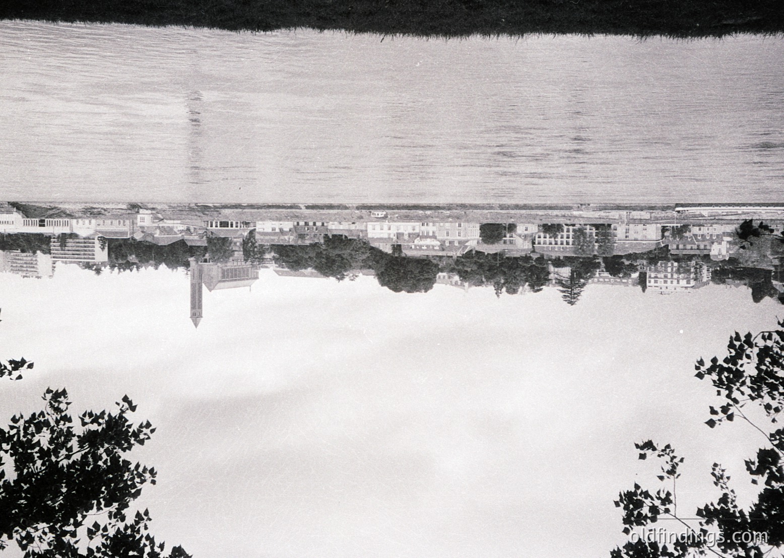 Aerial black-and-white photo of a coastal cityscape mirrored in calm waters, likely mid-20th century. Symmetrical reflection reveals linear streets, low-rise buildings, and a prominent pier extending into the sea. Vegetation frames edges, suggesting a seaside promenade or park.