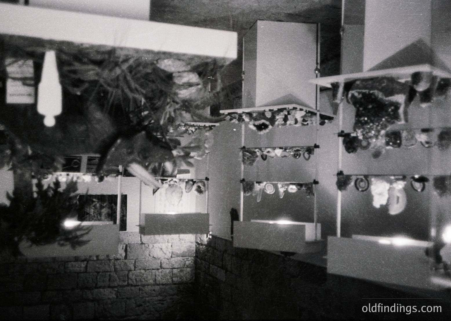 Black-and-white interior shot of a gallery or exhibition space featuring abstract geometric artworks. Framed pieces arranged in staggered rows on white panels against a dark brick wall, creating a dynamic visual rhythm. Minimalist design with sharp angles and contrasting textures. Likely mid-to-late 20th century (, ).