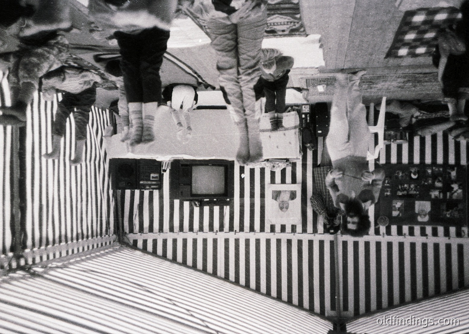 Vintage black-and-white photo of a playful indoor scene. Three individuals upside-down on striped bedsheets, suspended by a makeshift rig with a TV and wooden frame. Decor includes posters and a vintage radio. Likely 1960s–1970s, Western household setting.