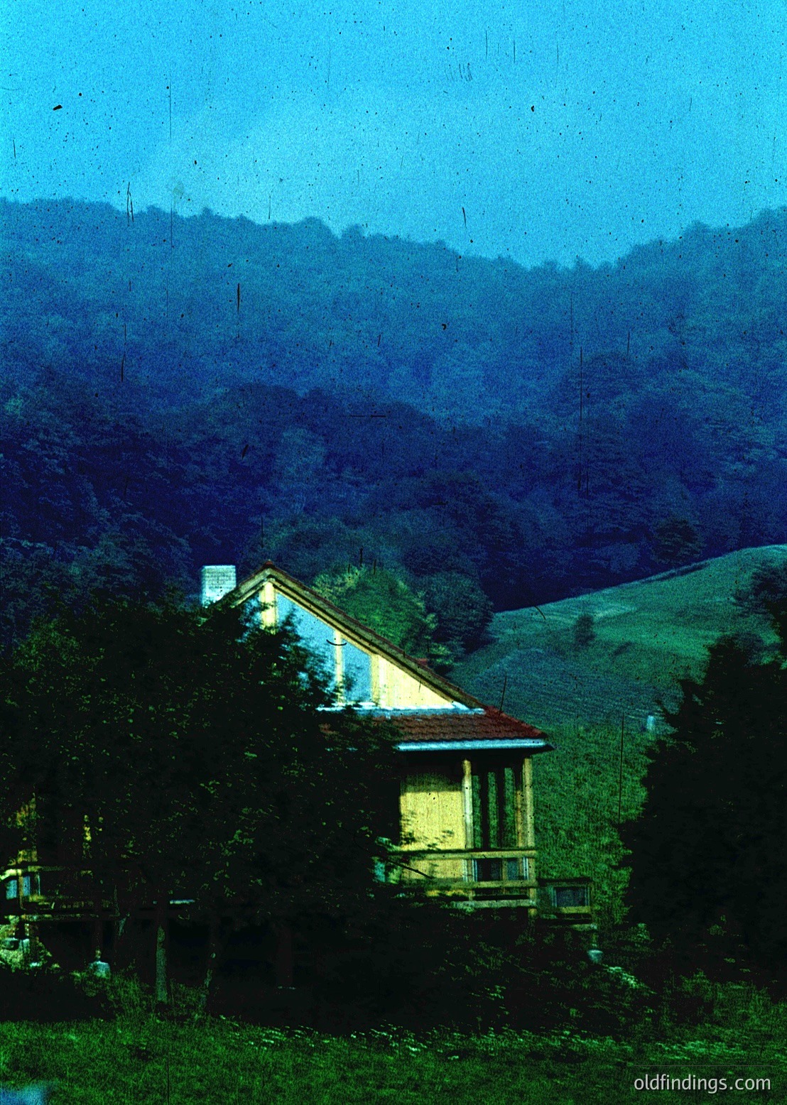 Vintage sepia-toned photo of a single-story house with a flat roof and balcony, nestled in a forested hillside. The structure features a chimney and large windows, surrounded by dense greenery. Likely mid-20th century European countryside.
