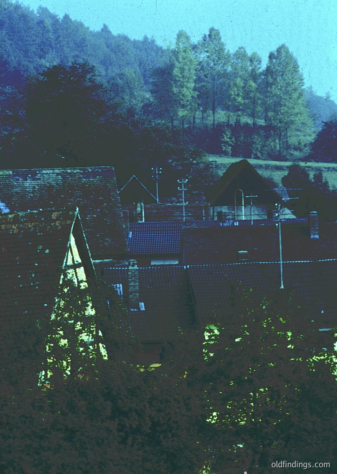 Aerial view of a rural village with distinct **traditional Bulgarian architecture**—notable for its **thatched roofs** and **brick chimneys**. The **tall church steeple** with a pointed cross suggests Eastern Orthodox influence. Surrounding **pine forests** and **rolling hills** indicate a mountainous or hilly region, likely in **southern Bulgaria**. The **1960s-1970s** era is suggested by the style and color tone.