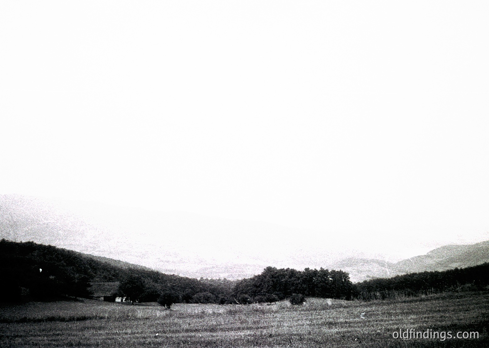 Black-and-white landscape featuring rolling hills, sparse trees, and a distant farmhouse. Low-contrast monochrome evokes mid-20th century rural photography.