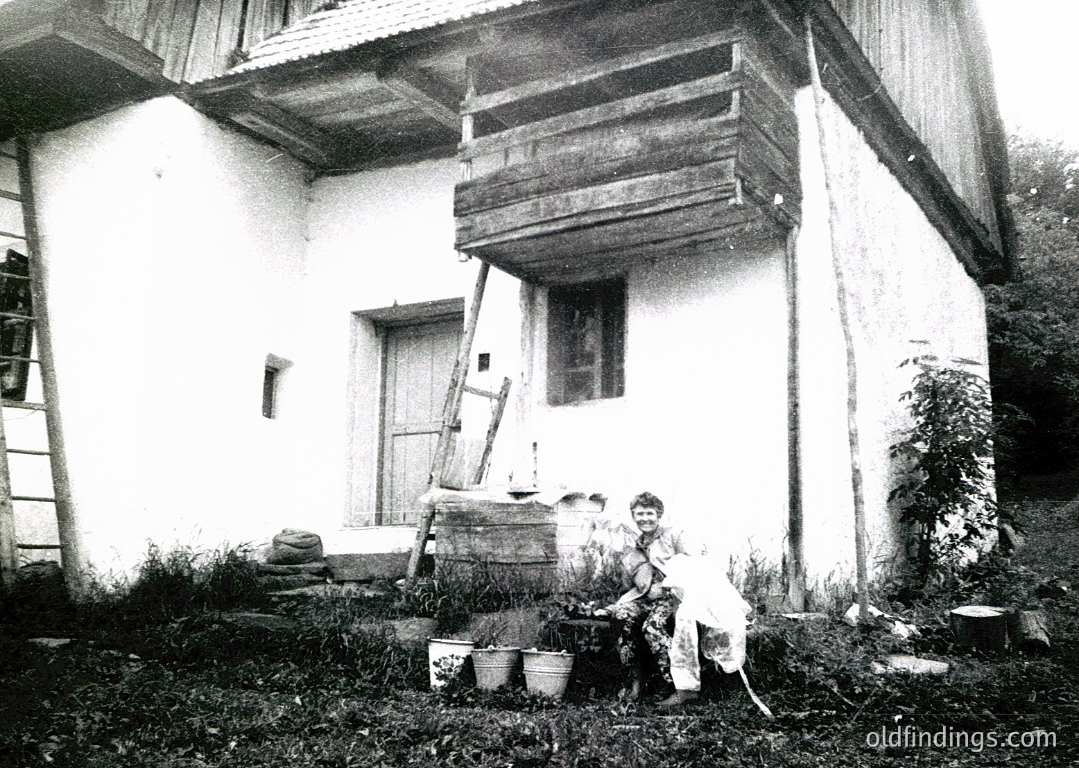 Traditional wooden house exterior with elevated balcony, likely rural Eastern Europe. Woman seated on steps, dressed in long-sleeve blouse and skirt, surrounded by clay pots and a wooden well bucket. Mid-20th century domestic life captured.