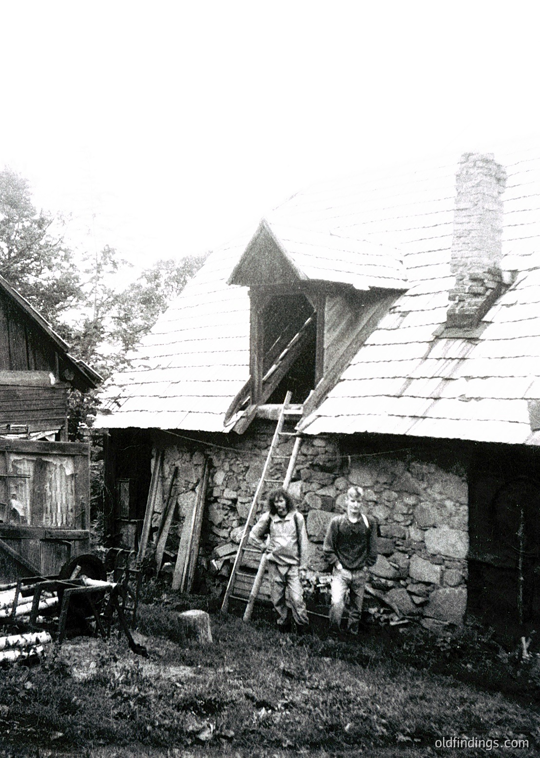 Rural stone cottage with wooden roof and exposed beams, likely Eastern European. Two men pose near a ladder and wooden cart, surrounded by agricultural tools. Stone chimney and rustic wooden door visible. Mid-20th century farming lifestyle.