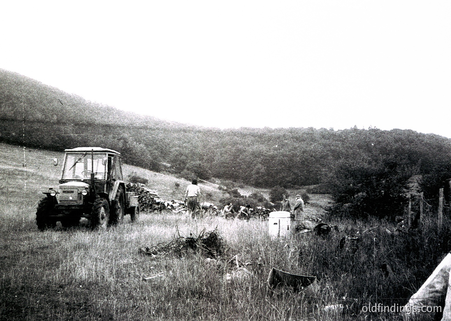 Vintage tractor hauling hay in an open field, surrounded by scattered wooden crates. Rolling hills and dense greenery in background. Mid-20th century rural farming scene.