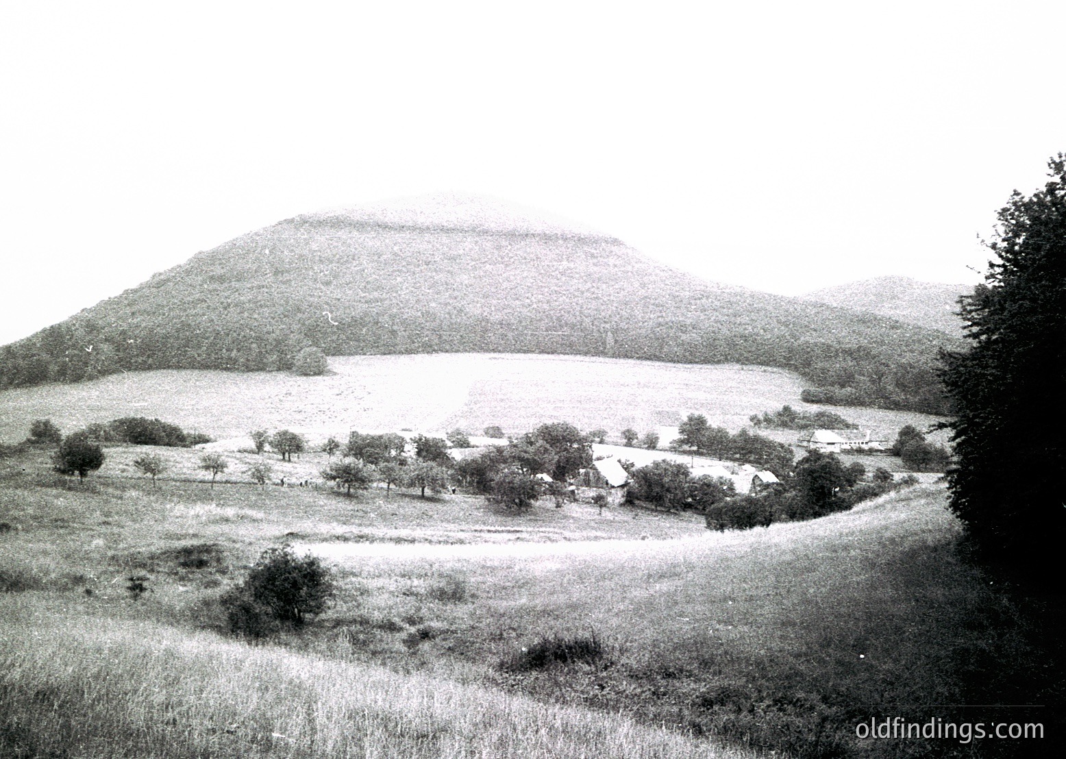 Vintage black-and-white landscape featuring a prominent artificial mound or hill, likely a burial tumulus, surrounded by open fields and sparse trees. Rural settlement with small structures visible at base. Mid-20th century agricultural or archaeological site.