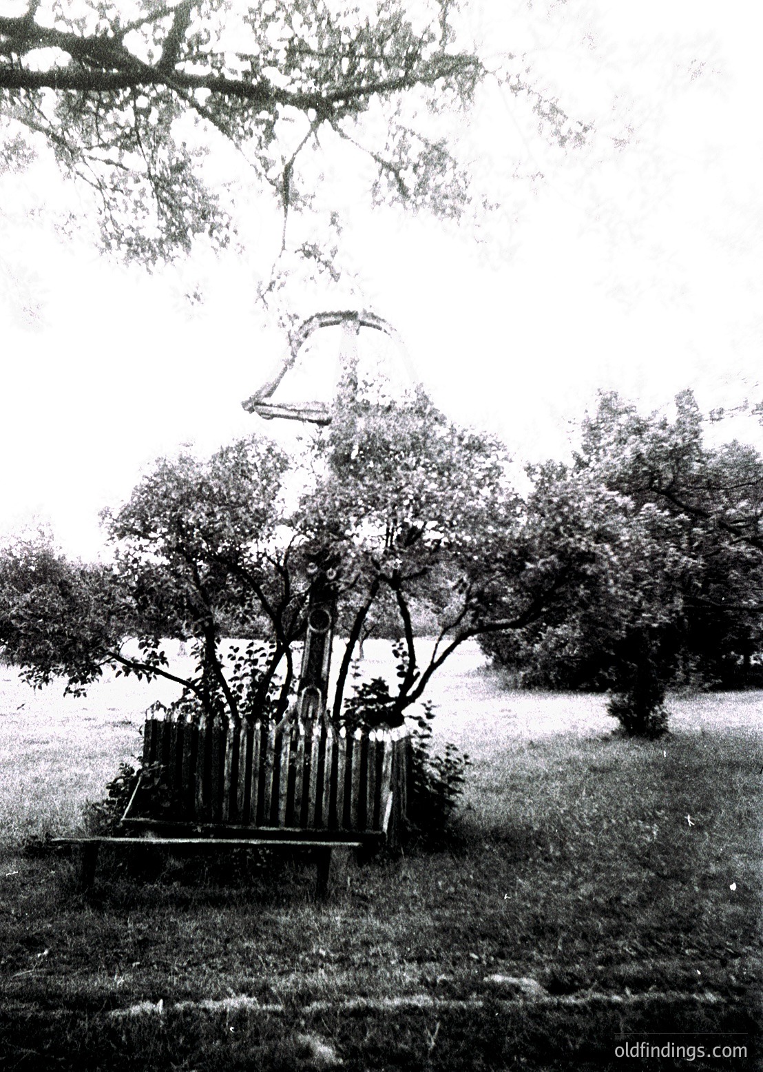 Vintage outdoor bell structure suspended from tree branches, surrounded by cylindrical concrete pillars. Likely a public park or garden feature, possibly from mid-20th century.