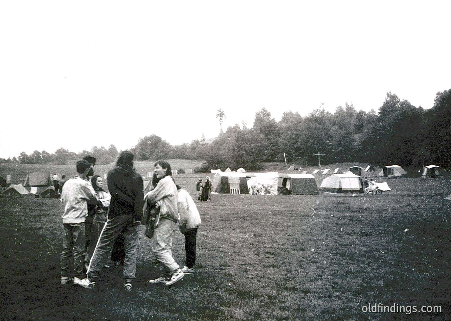 Group of four people in 1970s-style clothing—bell-bottoms, long sleeves, and layered scarves—standing in an open field surrounded by canvas tents. Wooden poles and makeshift structures suggest a temporary encampment. Dense forest in background indicates rural setting. Likely a festival or protest camp.